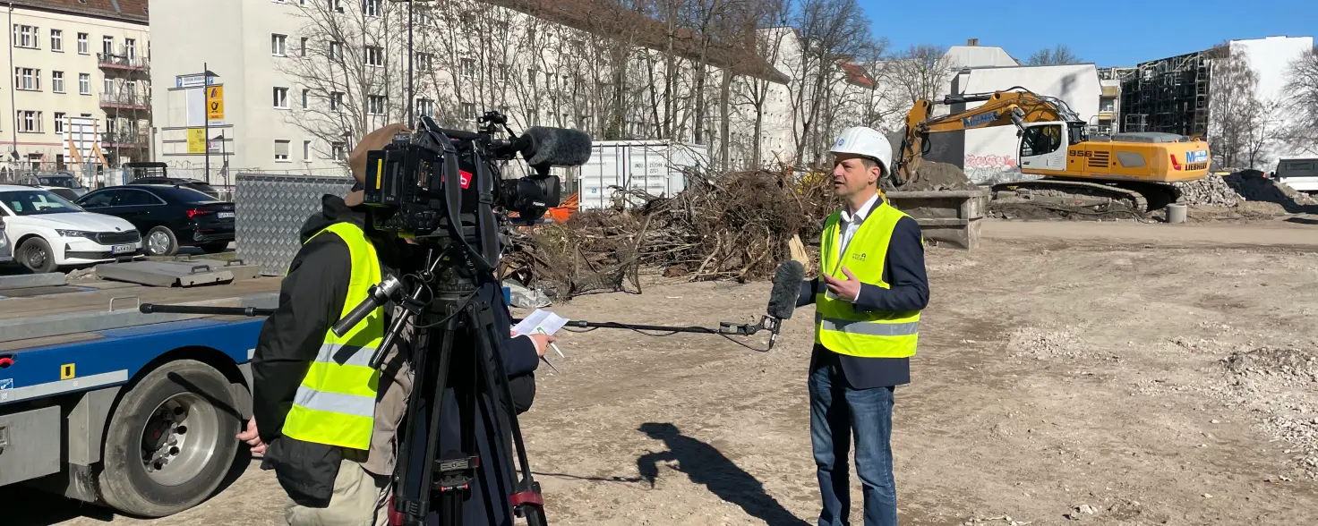 Ein Mann mit Helm und Weste wird auf einer Baustelle von der Presse interviewt
