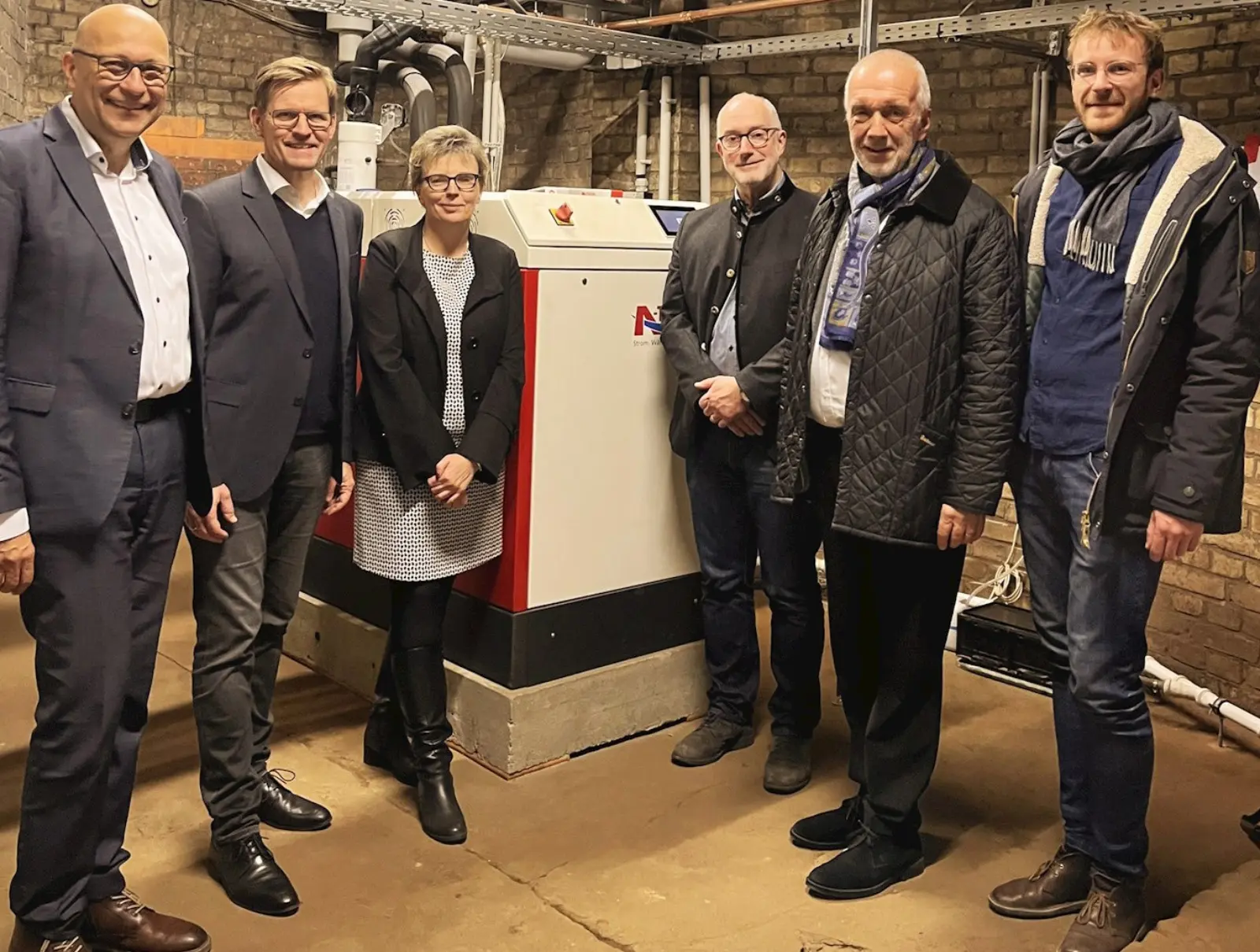 Projektbeteiligte stehen in der Energiezentrale der Apostel-Paulus-Kirche vor einem Blockheizkraftwerk zur Wärmeversorgung des Kirchengebäudes. Bild mit KI erstellt.