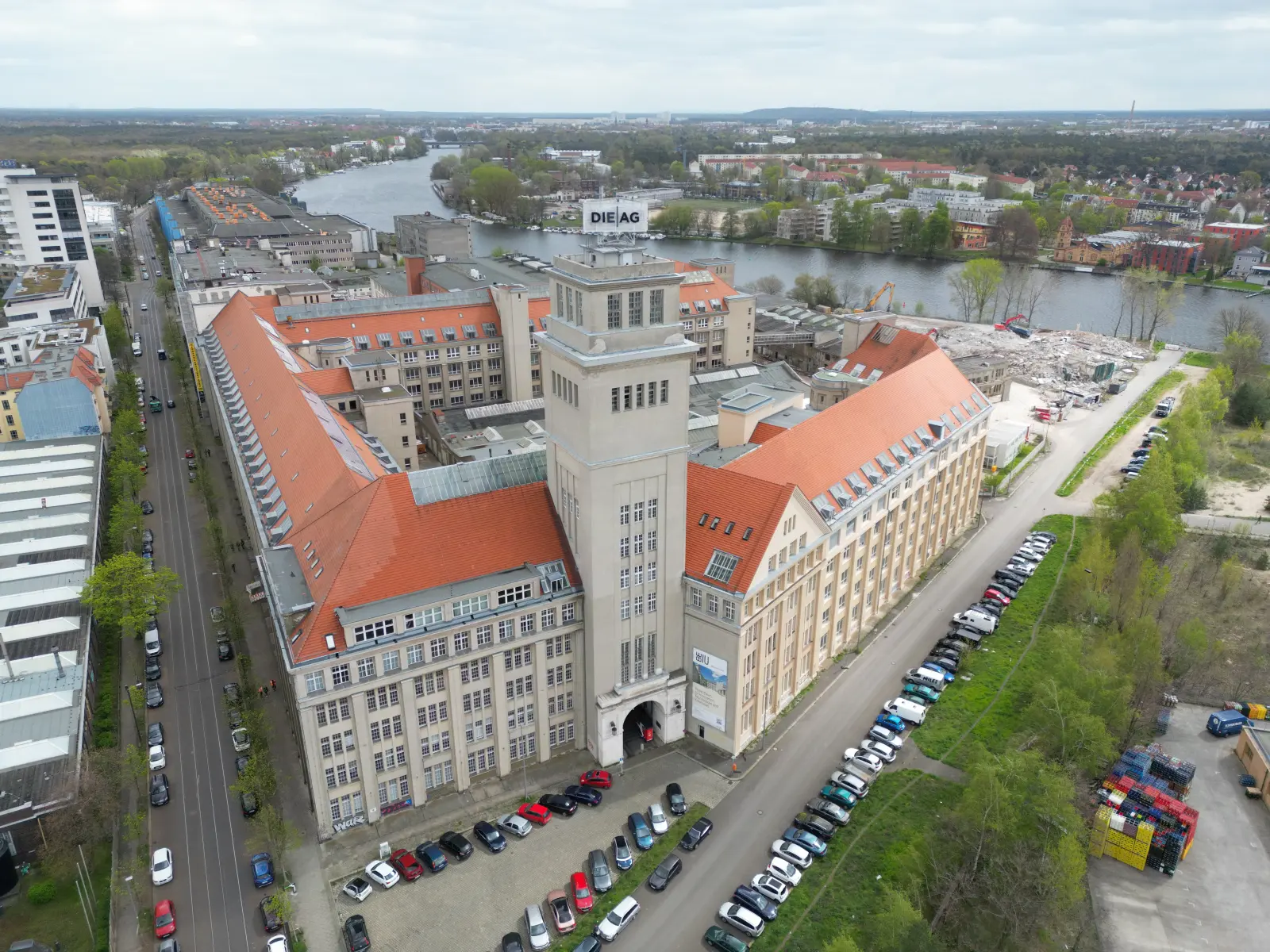 Luftaufnahme des historischen Behrensbaus am Behrens-Ufer in Berlin-Oberschöneweide mit markantem Uhrenturm und der Spree im Hintergrund. Bild mit KI erstellt.