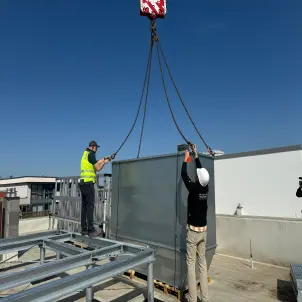 Fachkräfte montieren mithilfe eines Krans eine Einhausung für eine Wärmepumpenanlage auf einem Flachdach.