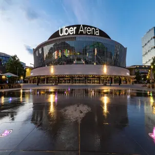 Uber Arena in Berlin bei Nacht mit beleuchtetem Schriftzug, Glasfassade und spiegelnden Wasserflächen auf dem Vorplatz. Bild mit KI erstellt.