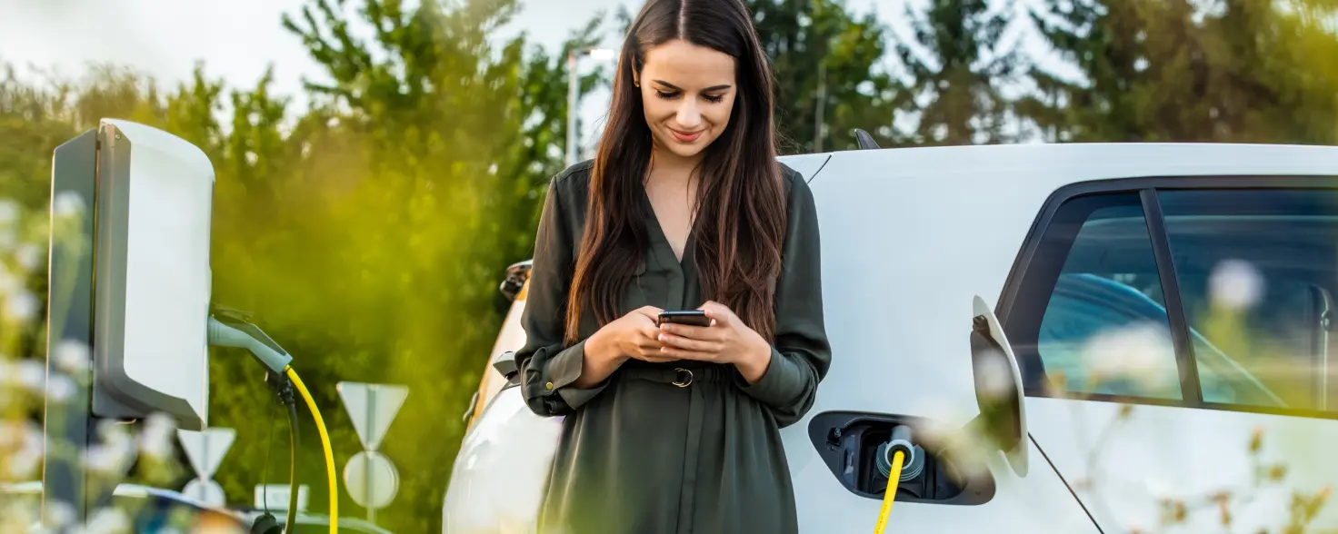 Eine Frau steht vor ihrem weißen E-Auto, das gerade geladen wird, und schaut lächelnd auf ihr Smartphone